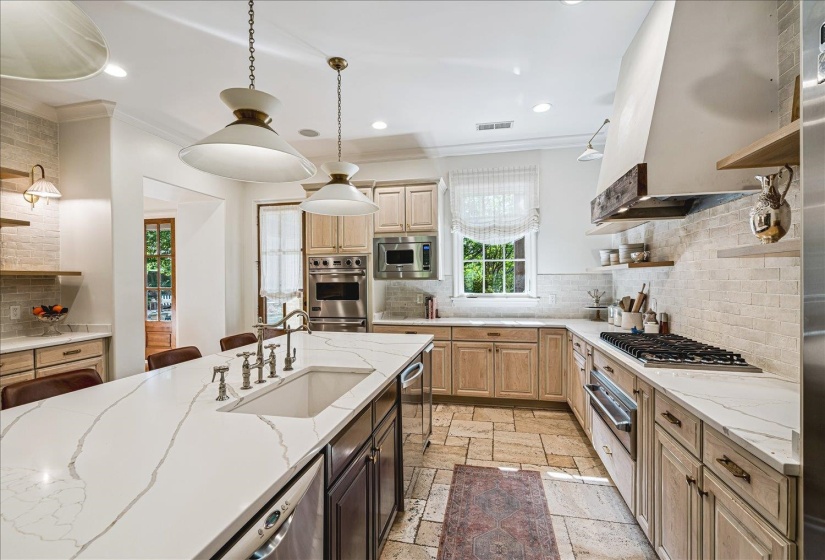 Kitchen featuring a large island with an integrated sink, light-toned cabinetry, light stone-finish flooring, a built-in oven and microwave, and a gas range with a custom range hood