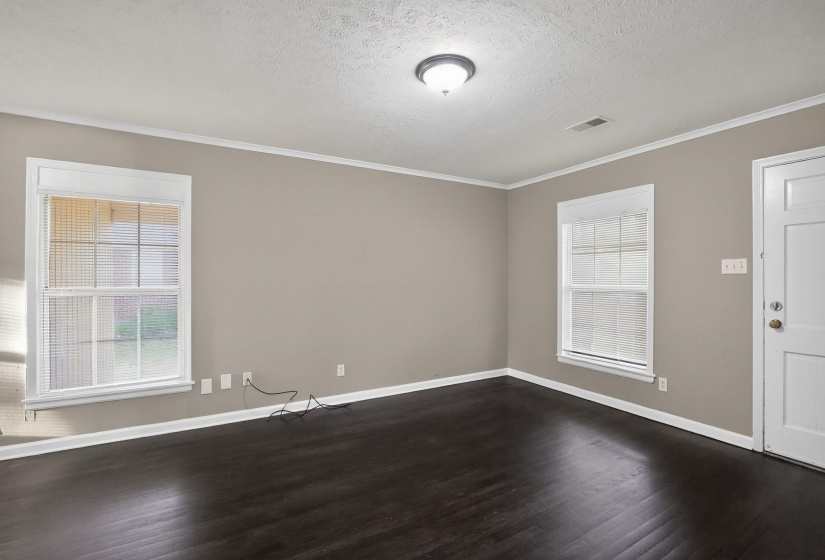 Spacious room featuring dark wood-finish flooring, two windows with blinds, a white paneled door, neutral wall paint, and white trim