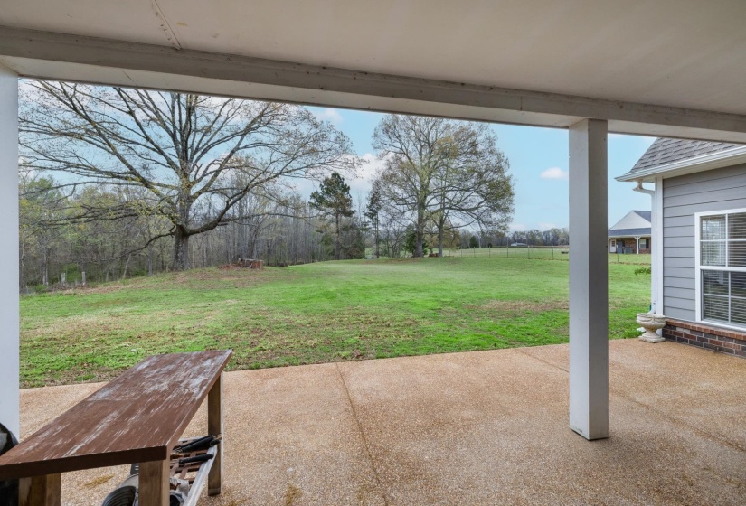Expansive covered patio featuring a textured concrete surface, white support columns, and views of a large grassy field with mature trees