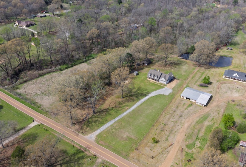 Expansive rural property featuring a main residence with a gray exterior and multiple gables, a long gravel driveway, a large barn structure with a metal roof, and a visible pond