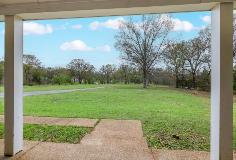 Covered porch with white support columns, concrete flooring, and an expansive grassy lawn with mature trees