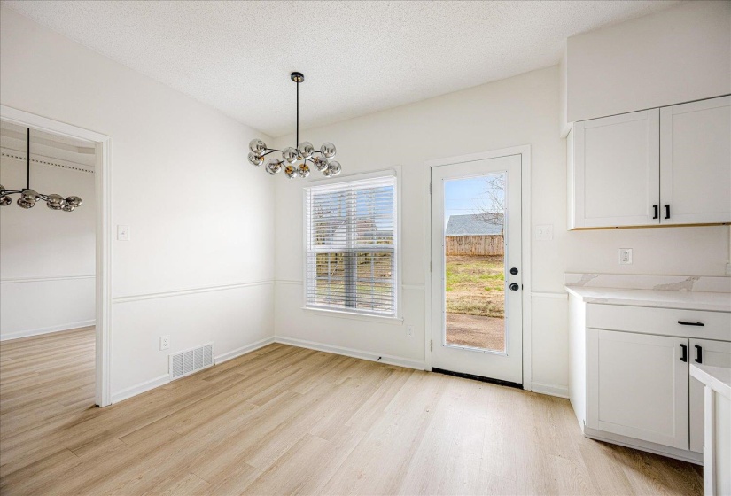 Dining area featuring wood-finish flooring, a modern multi-bulb chandelier, white shaker cabinetry with black hardware, a window with blinds, and an exterior door