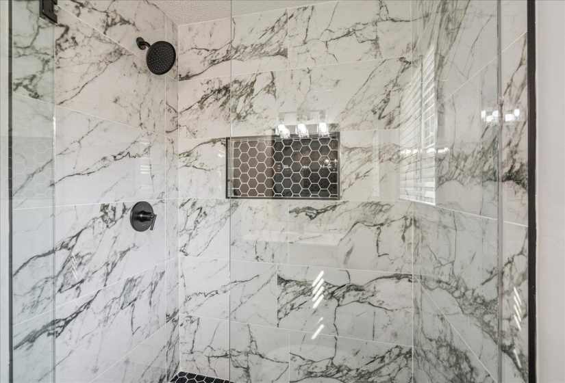 Shower enclosure featuring glass panels, large format marble-look tile, matte black fixtures, and a recessed niche with hexagonal mosaic tile