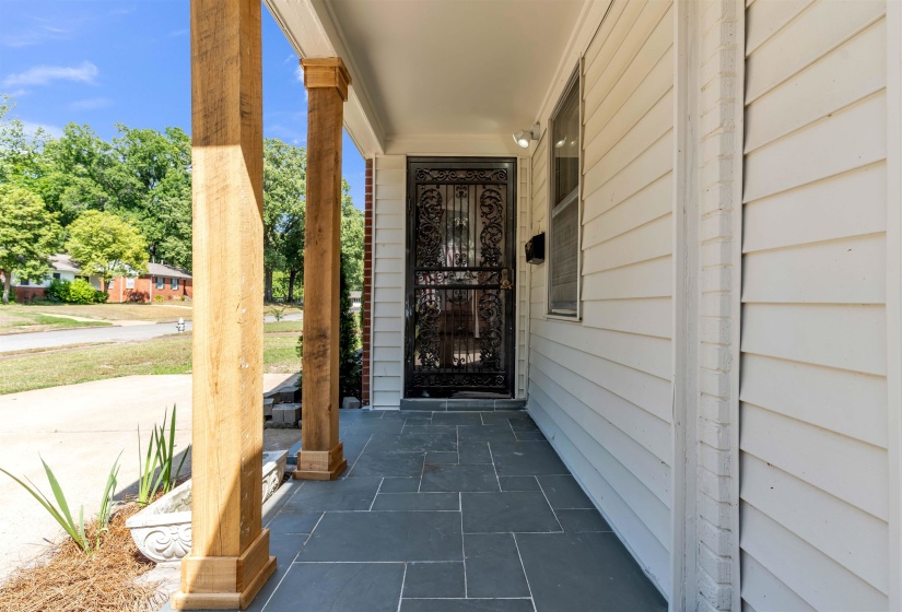 Covered front porch featuring natural wood columns, dark slate tile flooring, white siding, and an ornate black security door