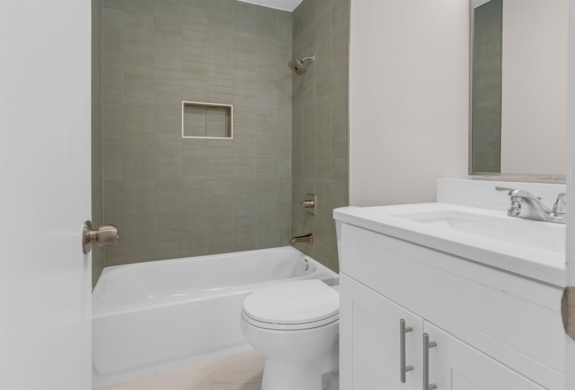 Bathroom featuring a white vanity with integrated sink, brushed nickel fixtures, a white commode, and a shower/tub combination with a tiled surround and recessed niche