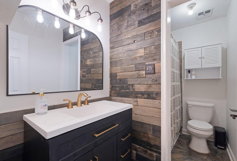 Bathroom featuring a vanity with a white countertop and gold-finish faucet, a rectangular mirror, a wall-mounted light fixture, and a wood-finish accent wall