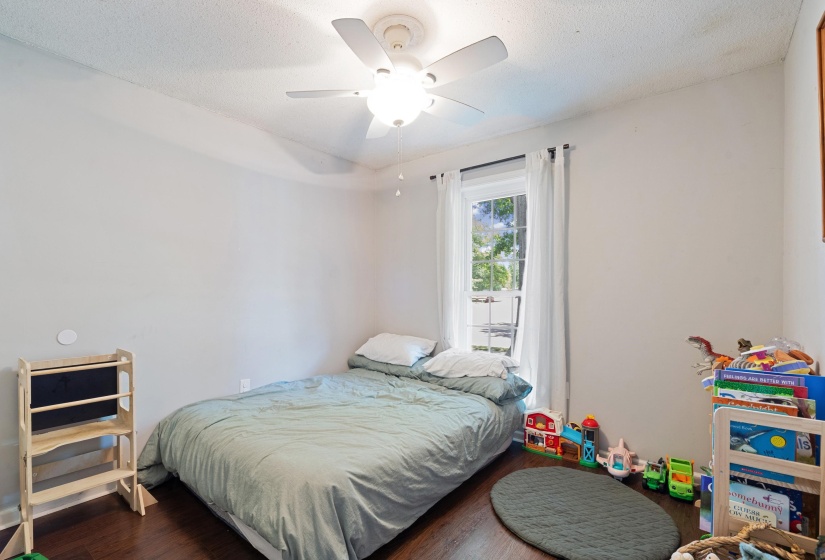 Bedroom featuring dark wood-finish flooring, a single window with white curtains, a ceiling fan with integrated lighting, and light-toned walls