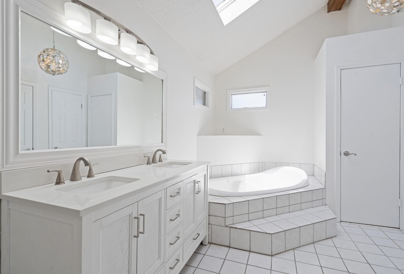 Spacious bathroom featuring a built-in tub with tiled surround, a large double vanity with brushed nickel fixtures, a skylight, and white tile flooring