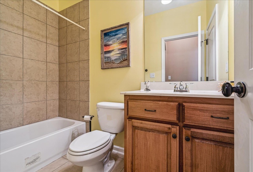 Bathroom featuring a vanity with wood-finish cabinetry and a white countertop, a built-in tub with neutral-toned tile surround, and a toilet.Located on first floor near second bedroom just off kitchen.