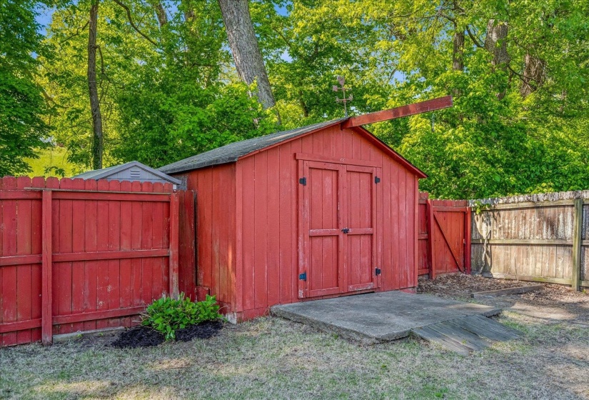 Red-painted storage shed with double doors, featuring a sloped asphalt shingle roof and a concrete pad base