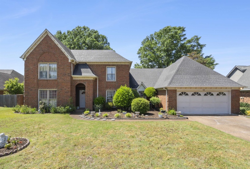 Brick facade residence featuring a covered entry, established landscaping, attached two-car garage, and a pitched shingle roof