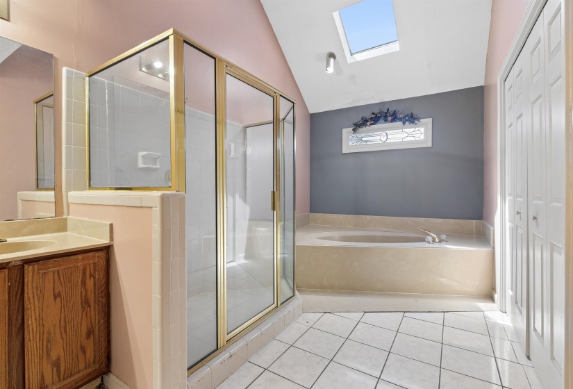 Spacious bathroom featuring a glass-enclosed shower, built-in soaking tub, a single vanity with wood-finish cabinetry, white tile flooring, and a skylight