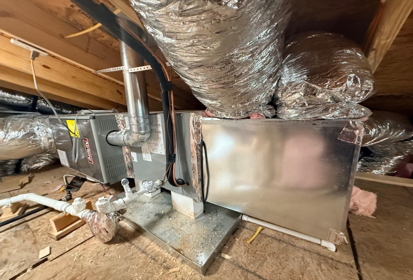 HVAC system featuring a furnace unit, insulated ductwork, and PVC piping, all situated on a plywood subfloor with exposed wooden ceiling joists