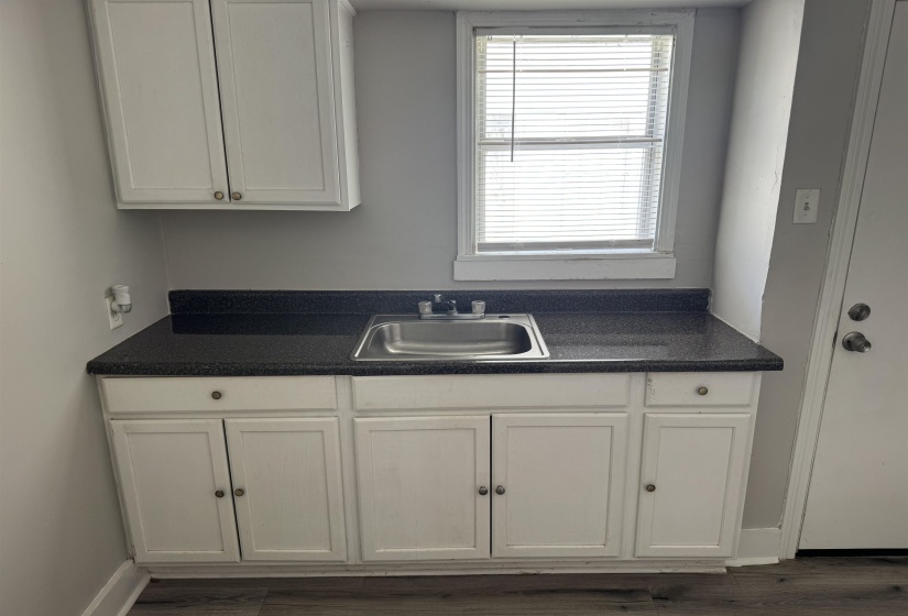 Utility area featuring a stainless steel sink, dark speckled countertop, white base and upper cabinetry, and wood-finish flooring
