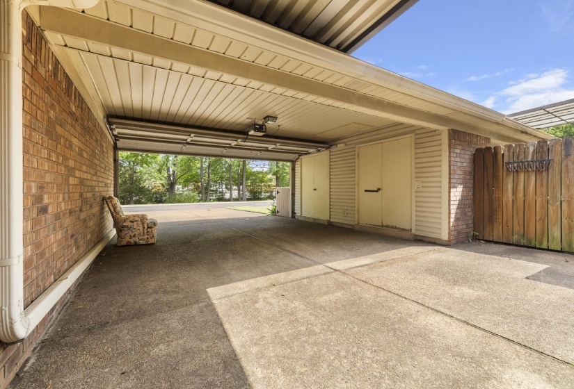 Covered carport with a concrete floor, featuring brick and siding walls, a wooden privacy fence, and integrated storage doors