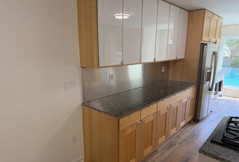 Kitchen area featuring light wood-finish lower cabinetry and upper cabinets with white high-gloss doors, contrasting granite countertops, and a stainless steel backsplash