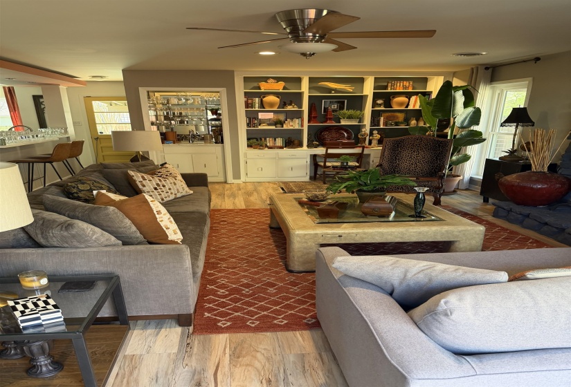Living area featuring light wood-finish flooring, a built-in bookshelf with lower cabinetry, recessed lighting, a ceiling fan, and a pass-through bar with pendant lighting