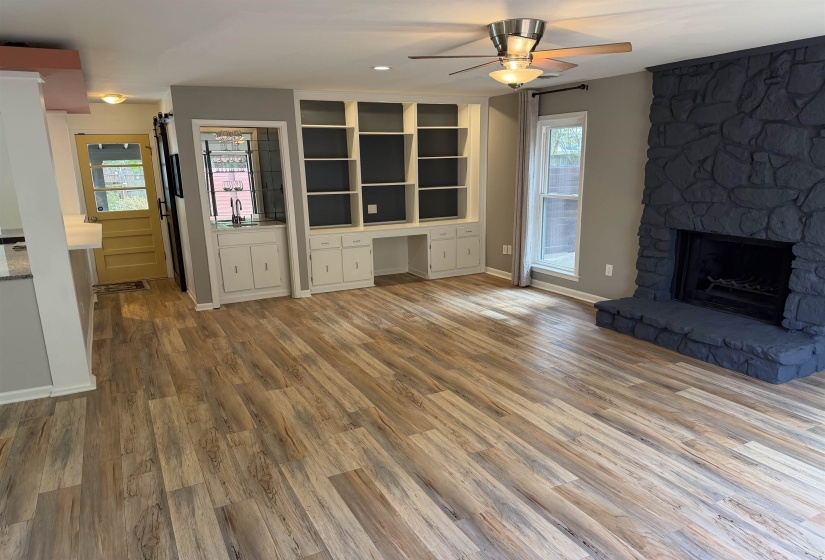 Spacious living area featuring plank wood-finish flooring, a dark stone fireplace, built-in shelving, a wet bar with a sink, and a ceiling fan with integrated lighting