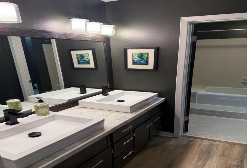 Dual vessel sinks with matte black fixtures, a mosaic tile countertop, dark wood-finish cabinetry, and wood-finish flooring