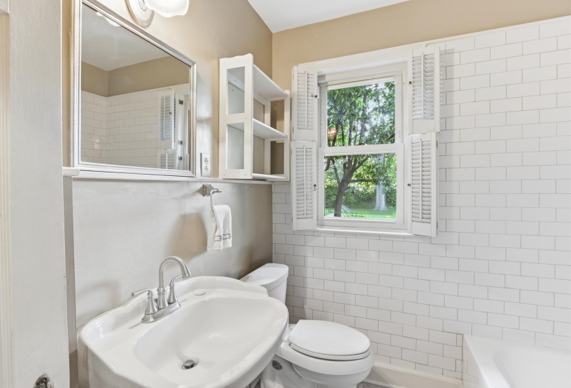 Bathroom featuring white subway tile walls, a porcelain pedestal sink with chrome fixtures, a window with louvered shutters, an overhead light fixture, and a built-in storage cabinet
