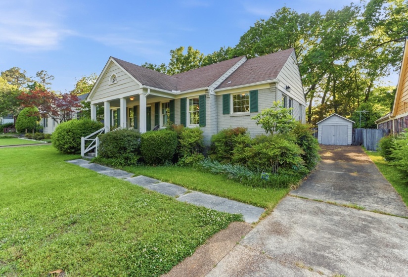 Traditional exterior featuring a covered front porch with white columns, light brick facade, green window shutters, a paved driveway, and a detached storage shed