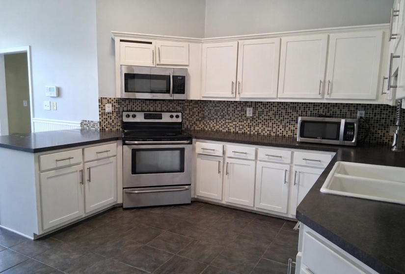 Open-concept kitchen featuring white cabinetry, dark countertops, stainless steel appliances, a mosaic tile backsplash, and tile flooring