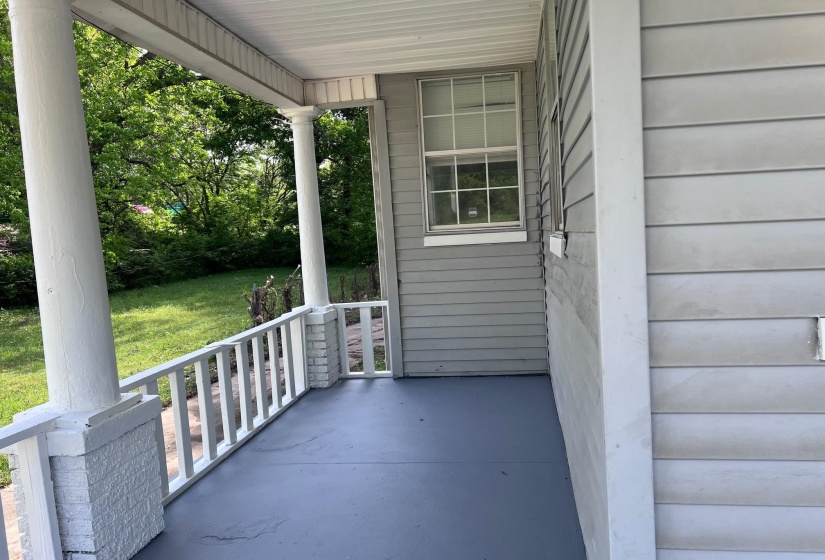 Covered front porch featuring a painted concrete floor, white spindle railing, and brick pilasters