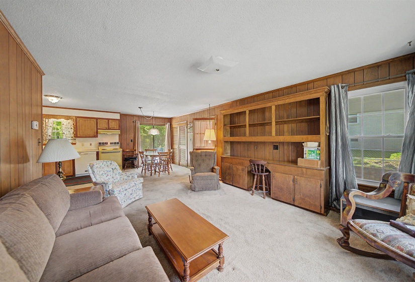 Living area featuring wood-paneled walls, built-in shelving, a large window, and carpeted flooring