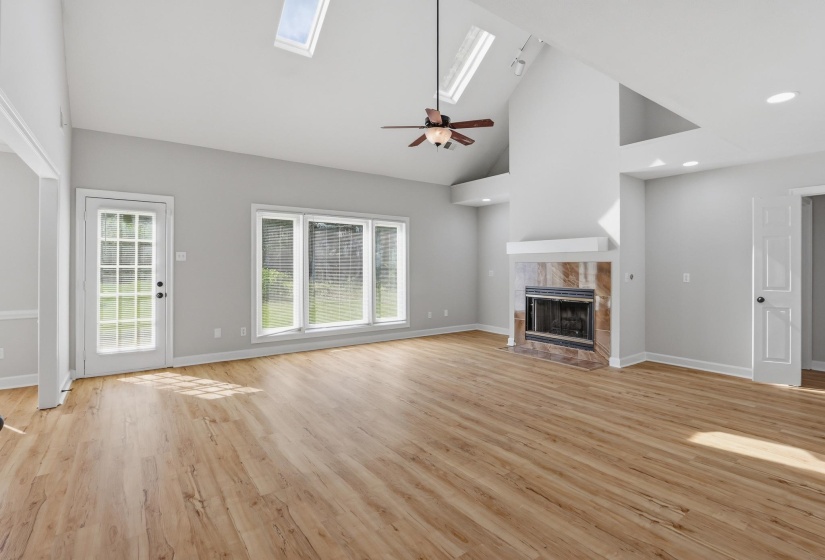 Bright living space featuring wood-finish flooring, a vaulted ceiling with skylights, a ceiling fan, and a fireplace with a stone surround