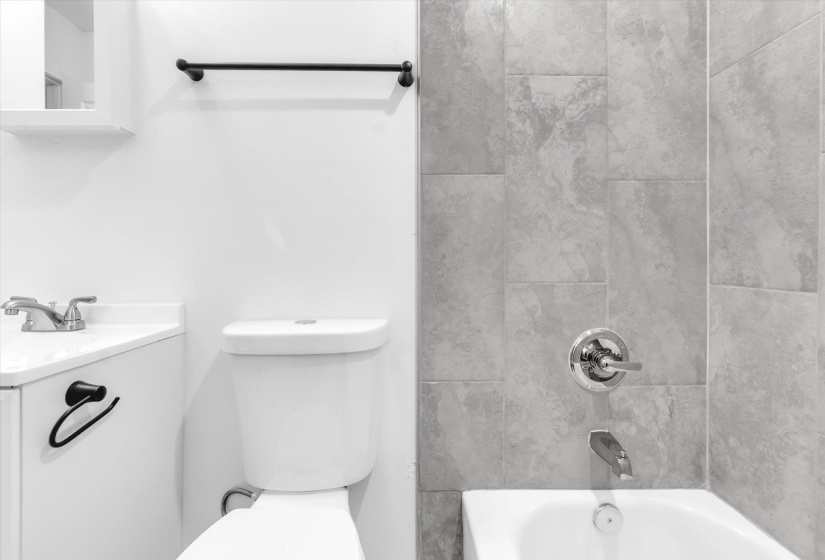 Bathroom featuring a white toilet, an integrated sink vanity with a white countertop, a black towel bar, a white wall-mounted cabinet, and large-format tile surrounding the bathtub