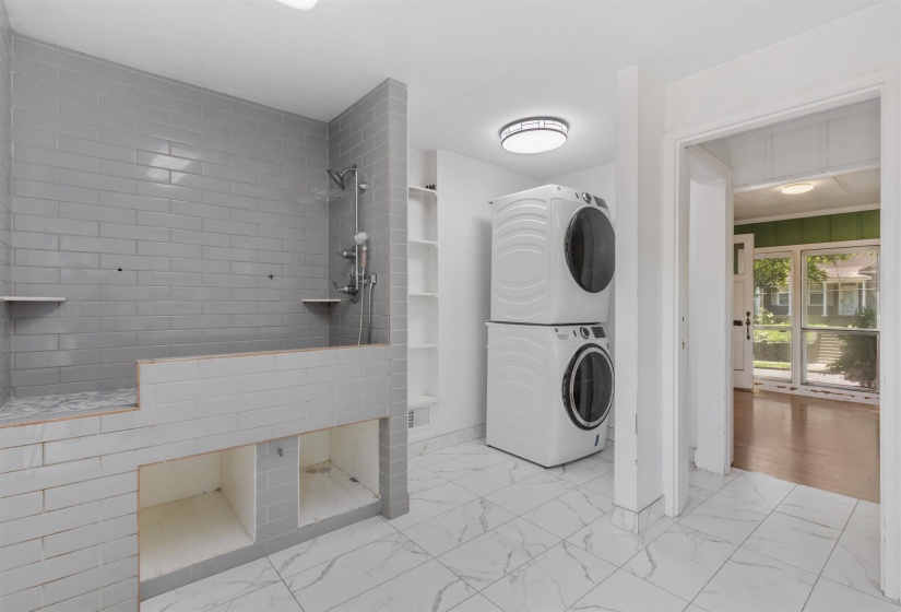 Tiled shower enclosure with built-in shelving, complemented by a stacked washer and dryer unit, white tile flooring, and integrated overhead lighting