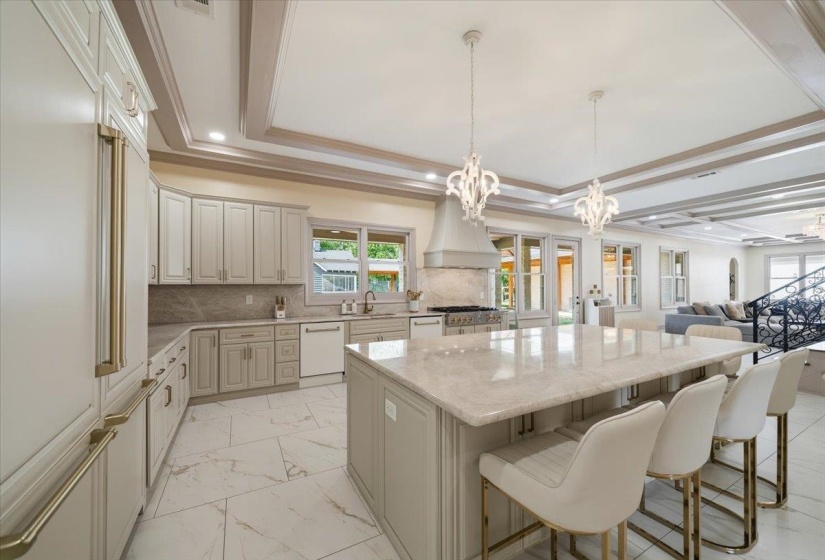 Gourmet kitchen featuring a large central island with seating, a professional-grade range with a custom range hood, extensive cabinetry with gold-toned hardware, stone countertops, and sophisticated coffered ceilings