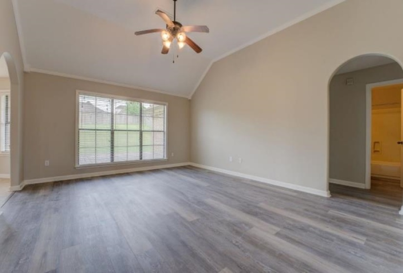 Spacious room featuring vaulted ceilings, wood-finish flooring, a large window with blinds, a ceiling fan with integrated lighting, and an arched doorway