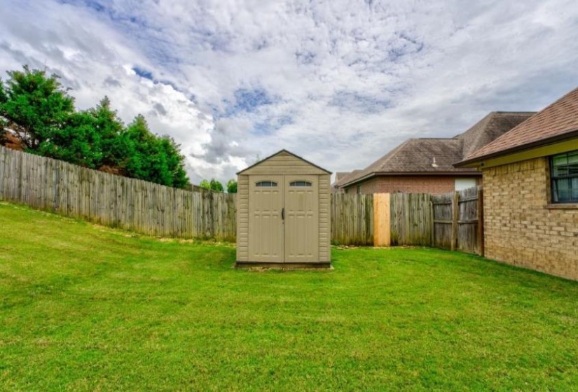 Expansive backyard featuring a manicured lawn, a light-colored storage shed, a wooden privacy fence, mature perimeter trees, and a brick exterior building with a brown shingle roof