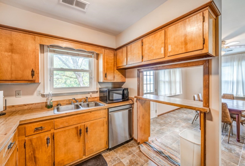 Kitchen featuring wood cabinetry, a double basin stainless steel sink, a stainless steel dishwasher, and a pass-through window with a breakfast bar