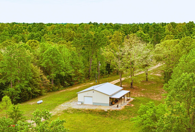 Metal-sided auxiliary building with a covered porch and white roll-up garage door, set amidst a dense wooded perimeter