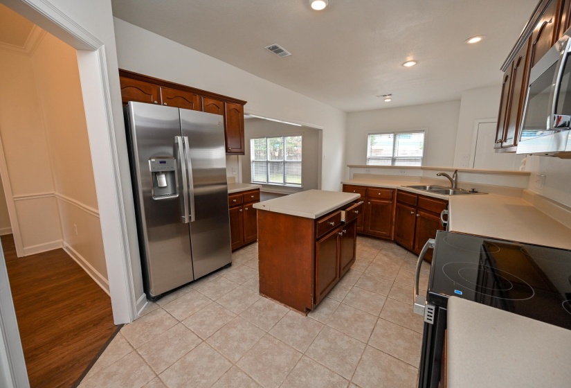 Spacious kitchen featuring an island, tile flooring, rich wood cabinetry, light-toned countertops, and stainless steel appliances including a refrigerator and built-in microwave