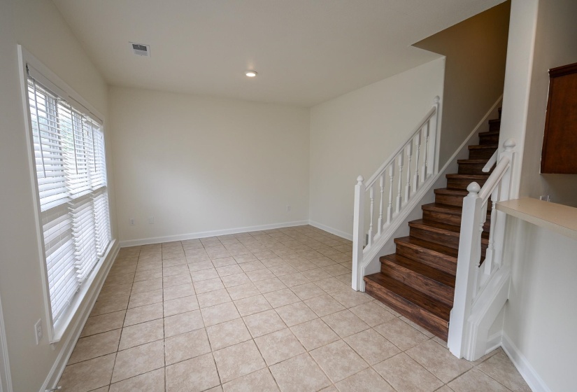 Spacious interior room featuring ceramic tile flooring, a prominent staircase with wood-finish treads and white balusters, and a large window with horizontal blinds