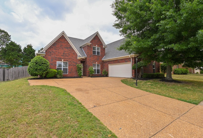 Traditional brick exterior featuring gabled rooflines, a two-car garage, mature landscaping, and an exposed aggregate driveway