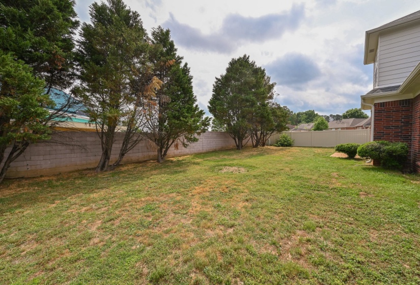 Expansive backyard featuring a green lawn, mature trees, a concrete block wall, and a white vinyl fence