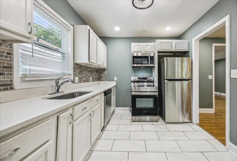 Modern kitchen featuring white cabinetry, light-toned countertops, a mosaic tile backsplash, stainless steel appliances, and large format floor tiling