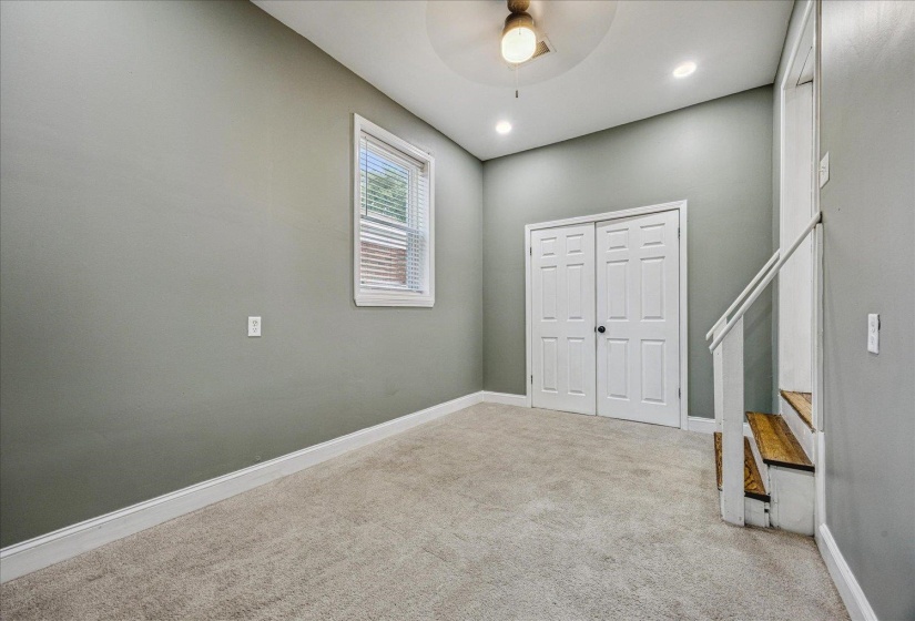 Carpeted room featuring a double-door closet, a single window with blinds, recessed lighting, and a partial staircase with wood-finish treads