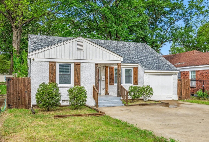 White stucco exterior featuring a gabled roof with gray shingles, wood-finish shutters, a single-car garage, concrete driveway, and mature landscaping