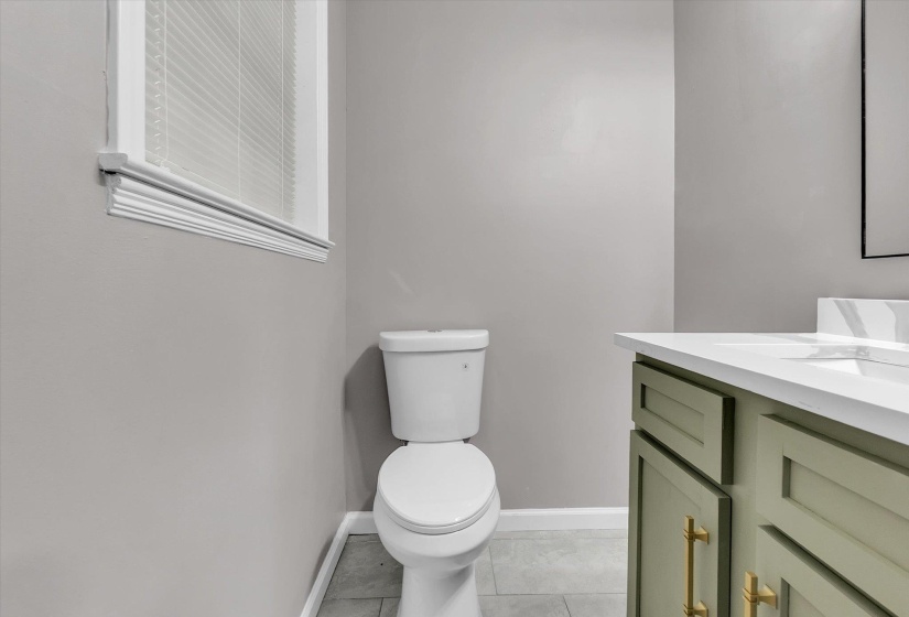 Bathroom featuring a white toilet, light gray tile flooring, a window with blinds, and a vanity with a white countertop and green cabinetry with gold-toned hardware