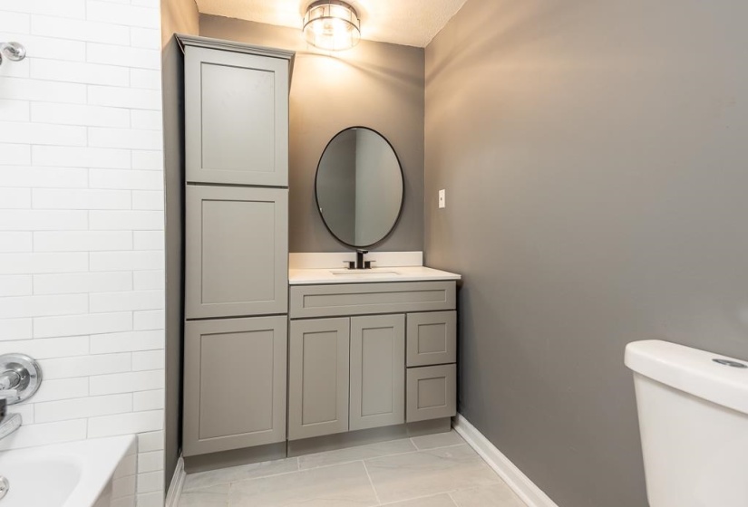 Bathroom featuring a gray vanity with a white countertop, a black-framed round mirror, and a tall matching storage cabinet