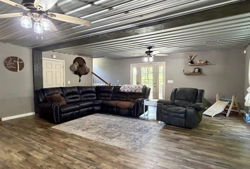 Spacious living area featuring wood-finish flooring, a corrugated metal ceiling with exposed beams, and ceiling fans with integrated lighting