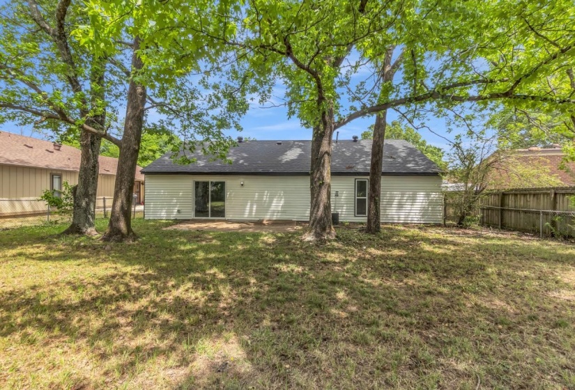 Spacious backyard featuring mature trees, a concrete patio, white horizontal siding, dark roof shingles, and sliding glass doors
