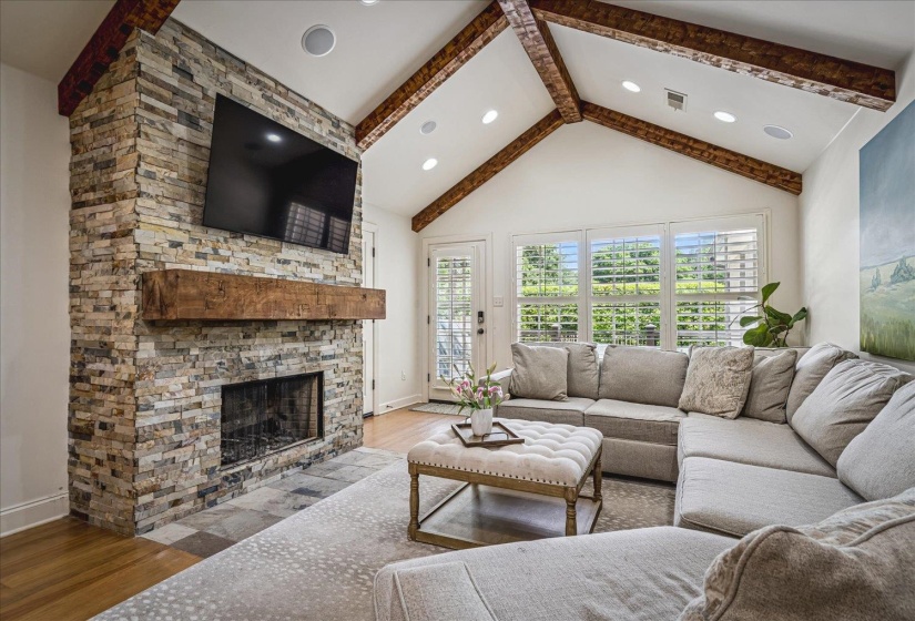 Vaulted ceiling with exposed wood beams, stone fireplace featuring a substantial wood mantel, wood-finish flooring, and white plantation shutters on multiple windows