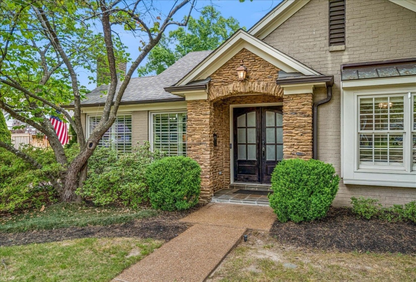 Stone-clad arched entryway with double doors, complemented by a bay window featuring white plantation shutters