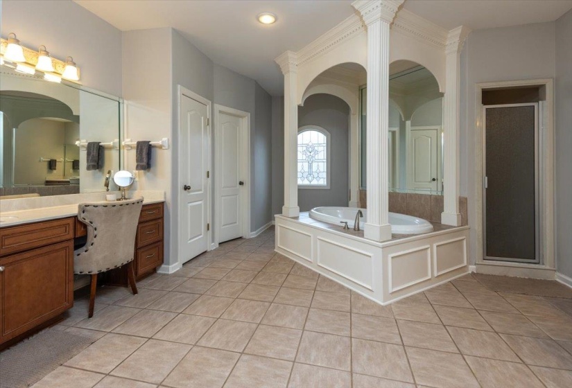Spacious bathroom featuring a built-in soaking tub with decorative columns, a glass-enclosed shower, a double vanity with ample counter space, and light-toned tile flooring